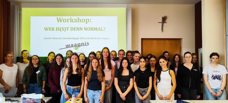 31 Schülerinnen und drei Erwachsene stehen in einem Klassenzimmer für ein Gruppenbild. Im Hintergrund läuft ein Beamerbild.